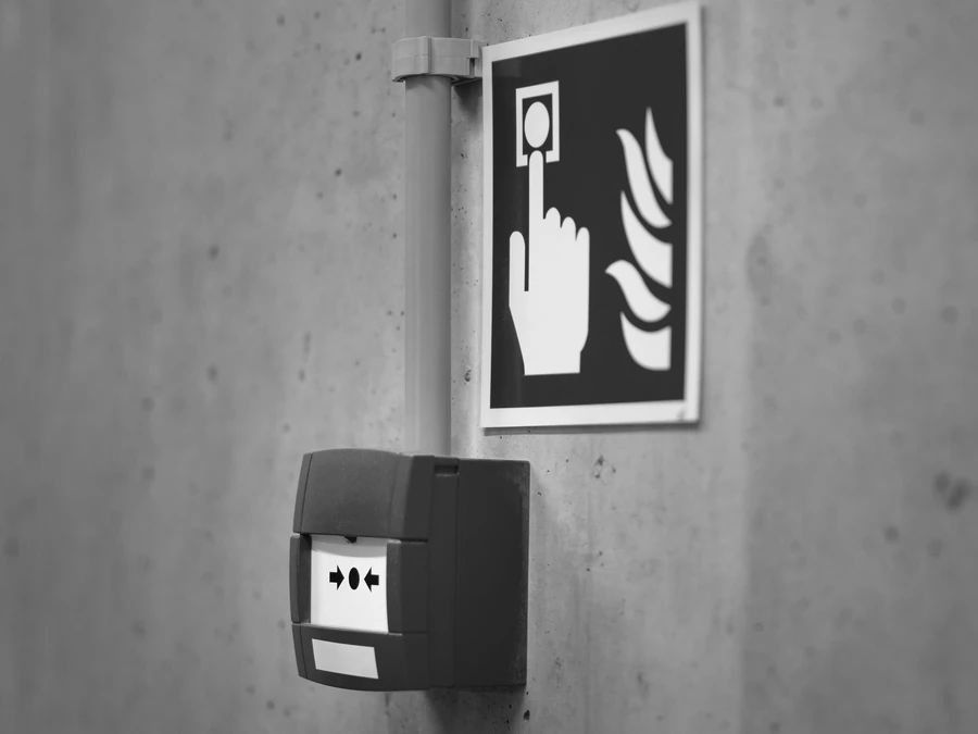 Pulsador de alarma contra incendios y cartel de señalización junto a una pared de hormigón, en blanco y negro